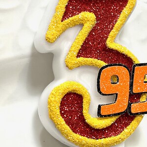 May include: A white birthday candle with the number 95 in orange glitter on a red and yellow glitter background.