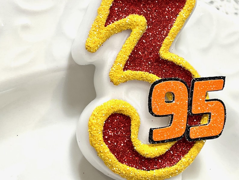May include: A white birthday candle with the number 95 in orange glitter on a red and yellow glitter background.