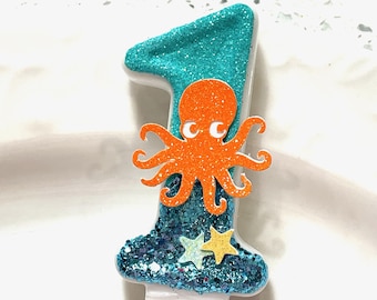 Può includere: Una candela di compleanno numero uno con la parte superiore e la base glitterate turchesi. Un polpo arancione è centrato sul numero. Due decorazioni a forma di stella sono alla base del numero. La candela è bianca.