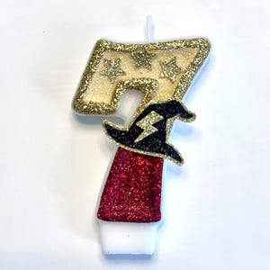May include: A number seven birthday candle featuring a gold glitter top section with three small stars, a red glitter bottom section, and a black glitter wizard hat accent with a white lightning bolt symbol centered on the front of the candle.