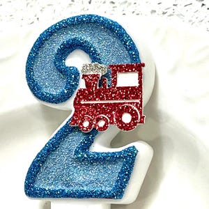 May include: A number 2 birthday candle with blue glitter and a red train decoration. The candle is white with blue glitter accents and a red train on top. The train has silver glitter accents.
