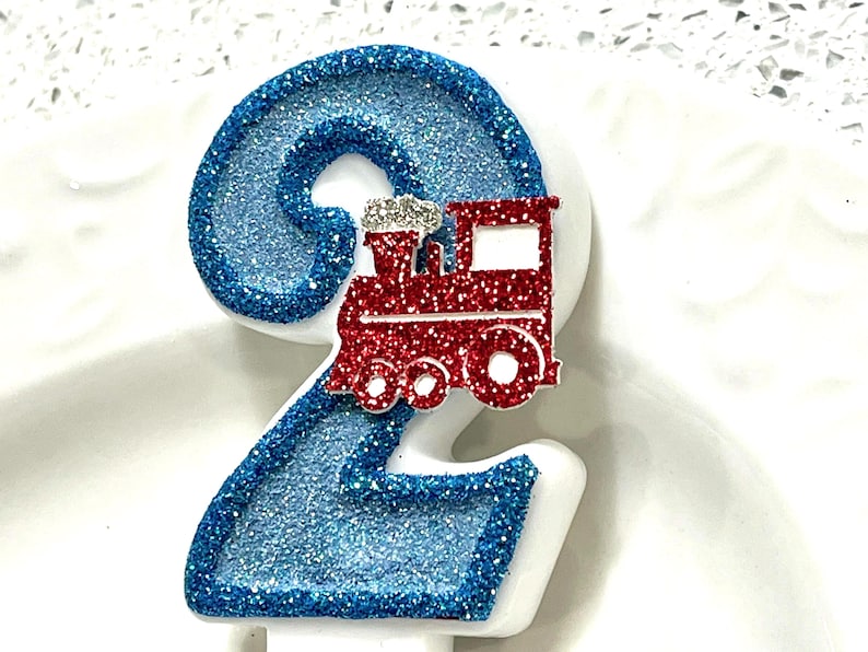 May include: A number 2 birthday candle with blue glitter and a red train decoration. The candle is white with blue glitter accents and a red train on top. The train has silver glitter accents.