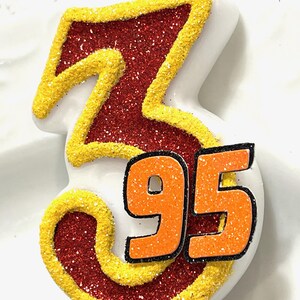 May include: A red and yellow glittery birthday candle shaped like the number 3 with the number 95 on the bottom.