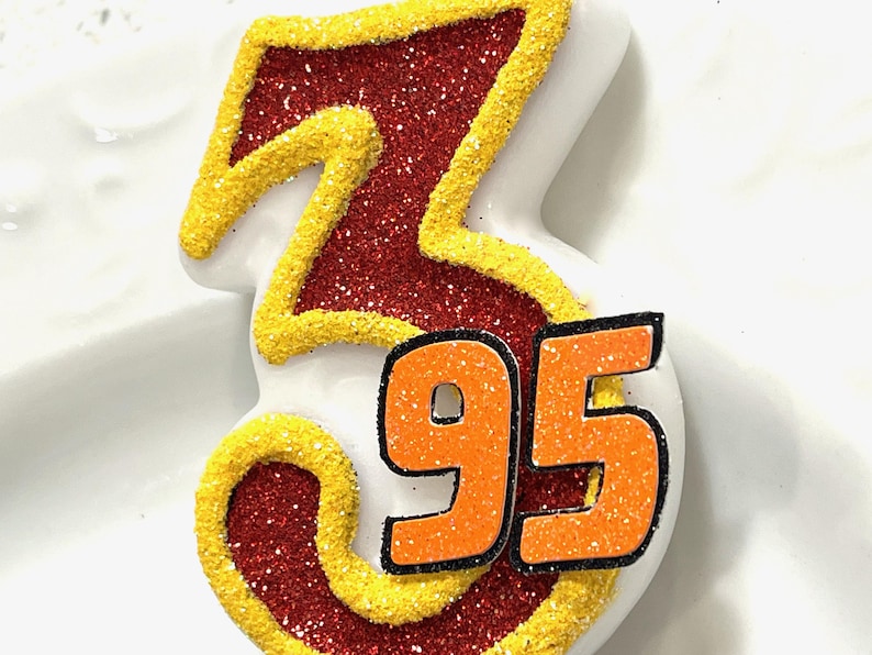 May include: A red and yellow glittery birthday candle shaped like the number 3 with the number 95 on the bottom.