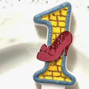 May include: A yellow and blue glittery number one candle with a red glitter shoe on the side. The shoe has a bow on the top.