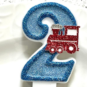 May include: A number 2 birthday candle with a red glitter train on top. The number is outlined in white and filled with blue glitter. The train has silver glitter accents.
