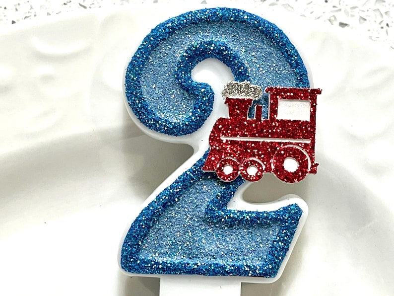 May include: A number 2 birthday candle with a red glitter train on top. The number is outlined in white and filled with blue glitter. The train has silver glitter accents.
