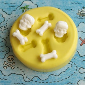 May include: A yellow silicone mold with skull and crossbones shapes. The mold contains white skull and bone shapes, perfect for crafting pirate-themed decorations. The mold is round and sits on a map-themed background.