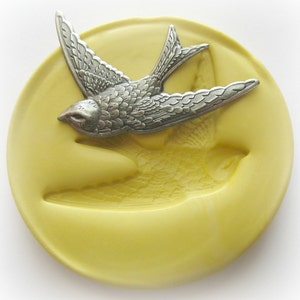 May include: A silver-colored bird-shaped mold sits on a yellow, circular base. The bird is detailed with textured feathers and a pointed beak. The base has an imprint of the bird's shape. The mold is likely for crafting or decorative purposes.