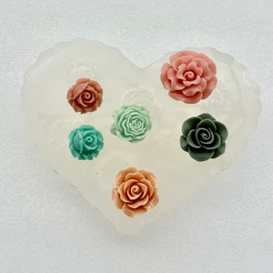 May include: A heart-shaped silicone mold with six rose-shaped embellishments in various colors: coral, teal, sage green, and dark green. The mold is translucent, and the roses are arranged in a circular pattern.