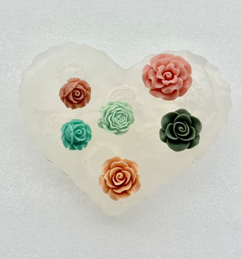 May include: A heart-shaped silicone mold with six rose-shaped embellishments in various colors: coral, teal, sage green, and dark green. The mold is translucent, and the roses are arranged in a circular pattern.