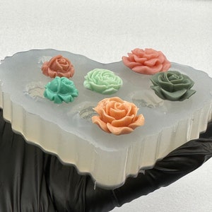 May include: A clear silicone mold holding six colorful rose-shaped soap pieces. The roses are in shades of peach, pink, green, and teal. The mold is held by a gloved hand.