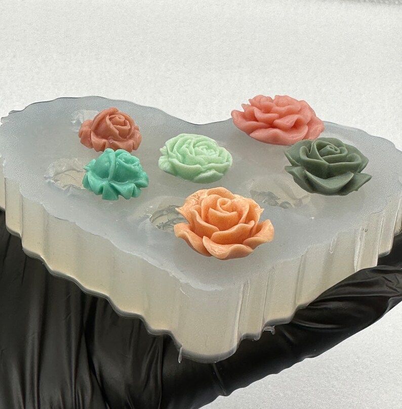 May include: A clear silicone mold holding six colorful rose-shaped soap pieces. The roses are in shades of peach, pink, green, and teal. The mold is held by a gloved hand.