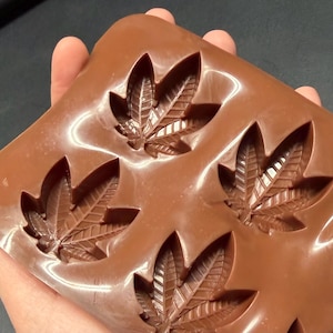 May include: A brown silicone mold with multiple leaf-shaped cavities. The mold is held in a hand and features detailed leaf vein patterns. The overall design suggests a theme related to cannabis or botanical elements.