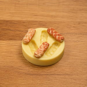 May include: A yellow silicone mold with three miniature bread loaves and impressions for more. The bread loaves are brown and tan, with detailed textures. The mold is circular and sits on a wooden surface.