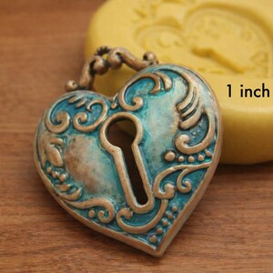 May include: A heart-shaped pendant, approximately 1 inch in size, with a keyhole design. The pendant features a teal and bronze color scheme with ornate scrollwork. A yellow mold is in the background.
