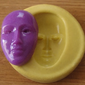 May include: A purple, glossy, face-shaped soap or resin casting next to a yellow mold with a face impression. The purple casting is a detailed replica of a human face. The yellow mold has a smooth surface and a face impression.