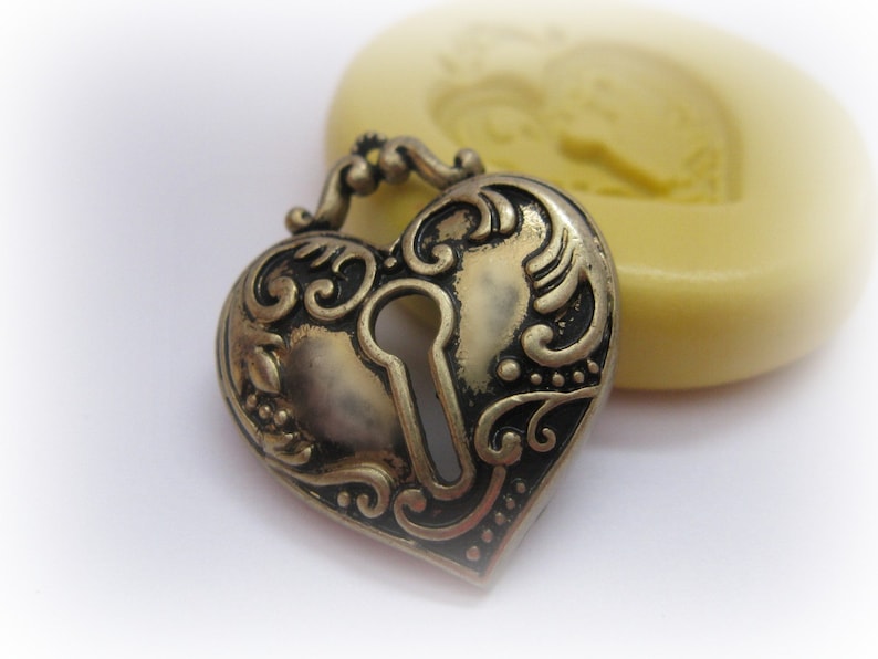 May include: A heart-shaped, antique-style locket with intricate scrollwork and a keyhole design. The locket is made of a bronze-colored metal and has a hinged clasp. A yellow, heart-shaped object is in the background.