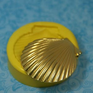 May include: A golden seashell-shaped mold and a gold seashell. The mold is yellow and round, with a seashell impression. The seashell is detailed with ridges and sits on top of the mold. The background is a blue patterned surface.