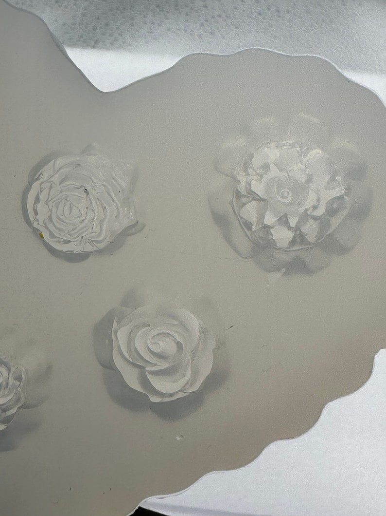 May include: A close-up shot of a clear, flexible mold with four raised floral designs. The flowers are white and appear to be roses. The mold has a wavy edge and is set against a white background.