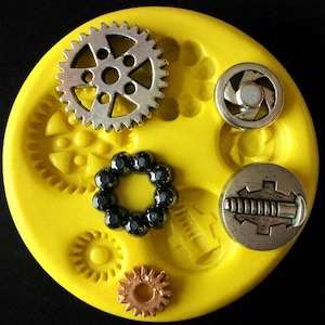 May include: A bright yellow silicone mold with various steampunk-themed embellishments. The mold features a large silver gear, a small copper gear, a black beaded ring, and two silver metal buttons. The background is black.