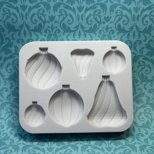 May include: A white silicone mold featuring six different Christmas ornament designs. The ornaments have various shapes, including round and bell-shaped, with detailed spiral and ribbed patterns. The mold is set against a teal patterned background.