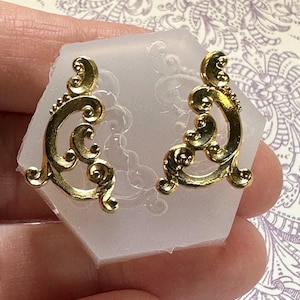May include: A pair of gold-colored, ornate earrings with a swirling, scroll-like design. The earrings are displayed on a clear, hexagonal mold. The background is a light purple floral pattern.