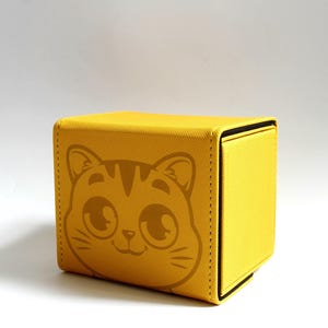 May include: A yellow, cube-shaped storage box with a cat face design on the front. The box is made of a textured material and has a magnetic closure. The cat face is a simple, cartoon-style illustration.