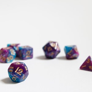 May include: A collection of colorful polyhedral dice, perfect for tabletop gaming. The dice feature a swirling mix of blue, purple, and gold colors. Various shapes are visible, including a D20, D12, D8, D6, and a D4. The numbers are in gold.