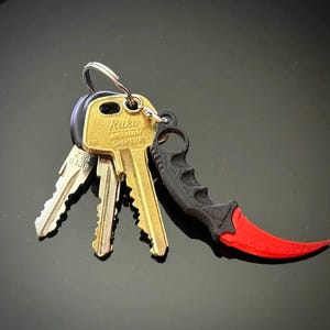 May include: A set of keys with a black and red karambit knife keyring. The keys are gold and silver, attached to a silver ring. The karambit knife has a black handle and a curved red blade.