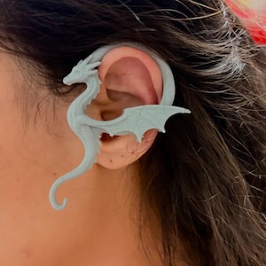 Puede incluir: Un pendiente de oreja de dragón gris claro. El dragón tiene alas detalladas y una cola curvada, diseñada para envolver la oreja. Este accesorio de fantasía es una pieza de joyería única.