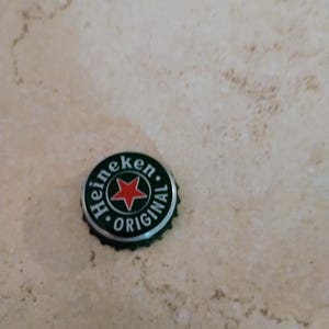 May include: A circular metal bottle cap with a dark green background, a central red star, and white text that reads Heineken ORIGINAL. The cap features a serrated edge and is designed for sealing a glass bottle, showcasing the brand logo clearly.