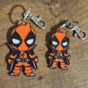 May include: Two orange and black Deadpool character keychains. The larger keychain is about 3 inches tall and the smaller keychain is about 2 inches tall.