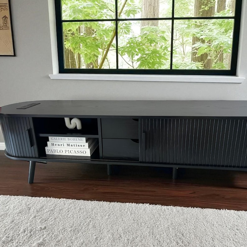May include: A black wooden media console with a curved design. It features a slatted door, drawers, and open shelving. Books with the titles "GALERIE BOHEME", "HENRI MATISSE", and "PABLO PICASSO" are visible on the shelf. A white sculpture sits on top.