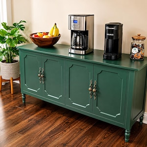 May include: A dark green cabinet with four doors, ornate handles, and a wooden countertop. A stainless steel coffee maker, a black coffee maker, and a glass jar of coffee pods sit on top. A wooden bowl of fruit is also present.