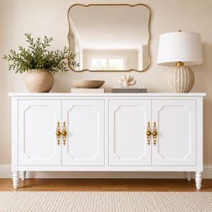 May include: A white cabinet with four doors and gold hardware. The cabinet is topped with a gold-framed mirror, a lamp, a vase with greenery, and decorative items. The cabinet has a classic design and is set against a neutral wall.