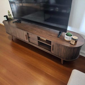 May include: A mid-century modern style TV stand in a warm brown finish. The stand features a slatted front design, two drawers, and open shelving. The stand is oval-shaped and has four tapered legs. A black TV sits on top.
