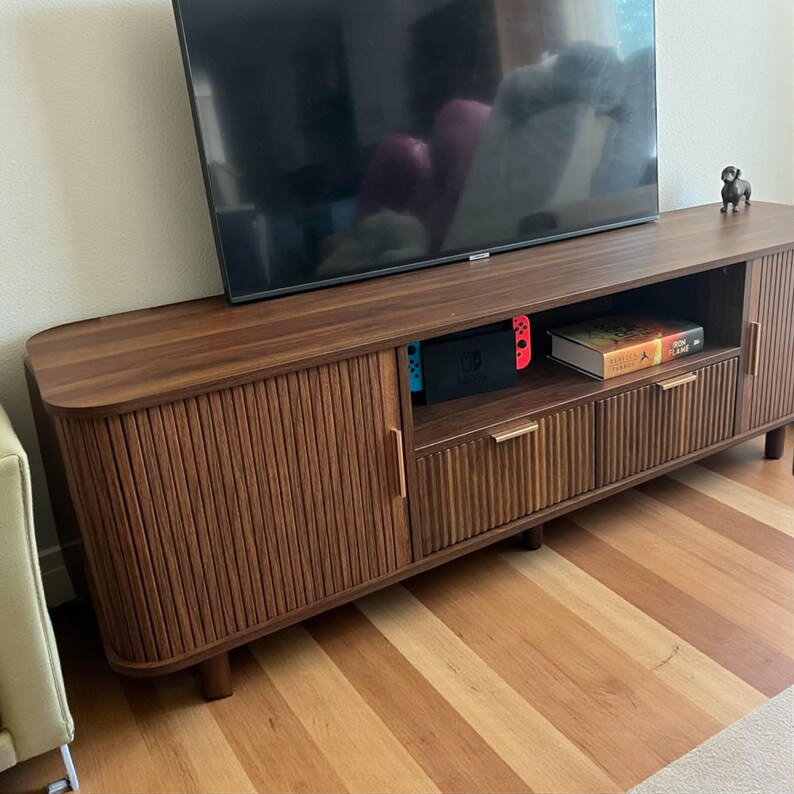 May include: A dark brown wooden TV stand with a large flat-screen television on top. The stand has two doors with vertical grooved detailing and two drawers. A Nintendo Switch and a book are visible inside the stand.