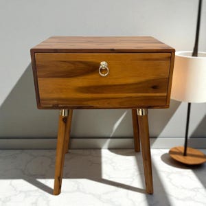 May include: A wooden nightstand with a single drawer and brass hardware. The nightstand has a warm, natural wood finish and tapered legs. The drawer features a brass ring pull. The nightstand is approximately 24 inches tall.