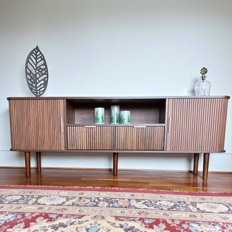 May include: A mid-century modern style wooden sideboard with vertical grooved doors and drawers. The cabinet has a dark brown finish and sits on tapered legs. Decorative items include a metal leaf sculpture, candles, and a glass decanter.