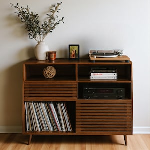 May include: A mid-century modern style record player console made of dark wood. The console has multiple compartments, including a section for vinyl records, a turntable, and a stereo receiver. Decorative items include a vase with greenery, a candle, and a framed photo.