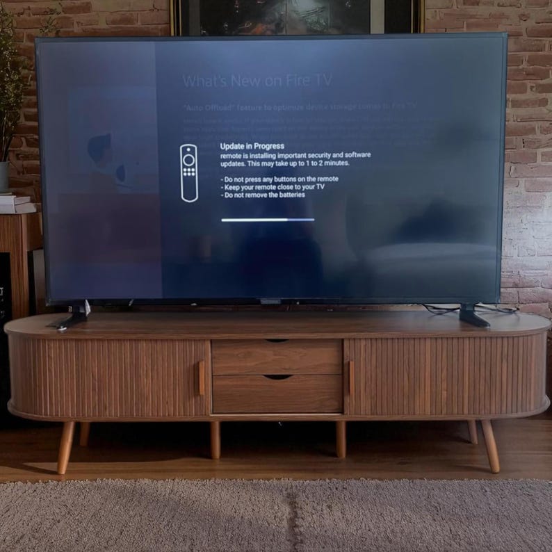 May include: A mid-century modern style TV stand in a warm brown finish. The stand has two doors with vertical grooves, two drawers, and tapered legs. A large flat-screen TV displays the text "What's New on Fire TV" and an update in progress message.