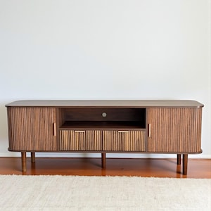 May include: A mid-century modern style TV stand in a warm brown finish. The stand features two doors with vertical ribbed detailing, a central open shelf, and a drawer with two compartments. It stands on four tapered legs.