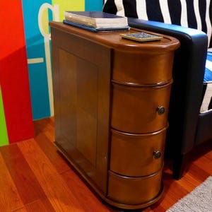May include: A narrow, wooden side table with three drawers and a curved front. The table is a warm brown color and has a small, rounded top. A book and a coaster sit on top of the table. The table is next to a black couch.