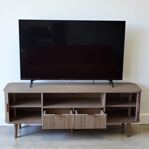 May include: A light wood-tone TV stand with a large black television on top. The stand has open shelving, two drawers with vertical ribbed detailing, and wooden legs. The drawers have brown leather-look handles.