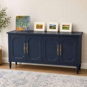 May include: A navy blue sideboard with four doors and brass hardware. Three framed landscape paintings and a decorative plate with a floral design rest on top. The sideboard has a traditional design and is set on a patterned rug.