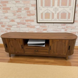 May include: A mid-century modern style wooden TV stand with a curved top and legs. The stand has two doors with vertical grooved panels, a drawer, and an open shelf. A white game console sits on the shelf.