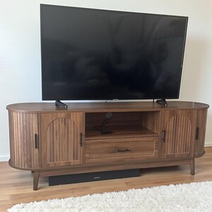 May include: A wooden TV stand with a dark finish, featuring two cabinet doors with vertical grooves and a central drawer. The stand has a mid-century modern design with curved edges and black hardware. A large black television sits on top.