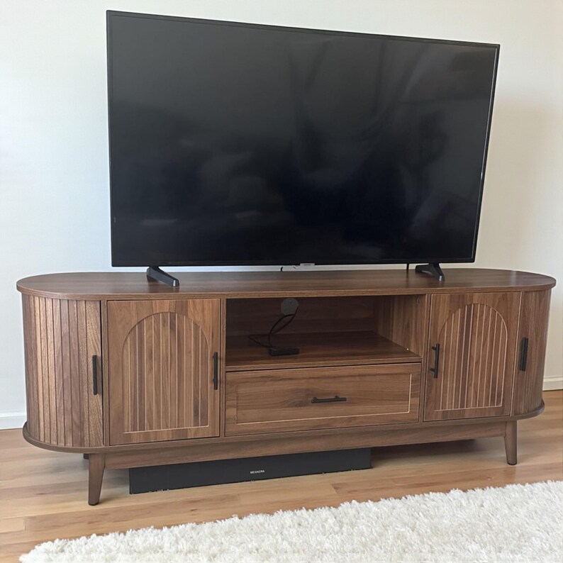 May include: A wooden TV stand with a dark finish, featuring two cabinet doors with vertical grooves and a central drawer. The stand has a mid-century modern design with curved edges and black hardware. A large black television sits on top.