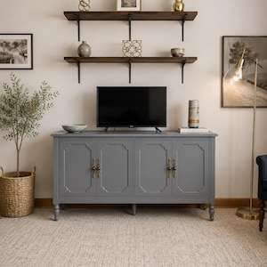 May include: A gray cabinet with four doors and ornate brass handles. A flat-screen TV sits on top of the cabinet. Two dark wooden shelves with decorative items are mounted on the wall above the cabinet.